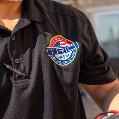 Close-up of a person wearing a Trio Heating Air Plumbing logo shirt, pen in pocket, working with tools.